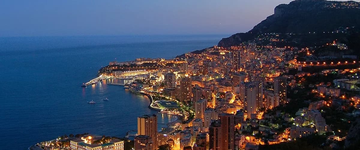 Night panoramic view of the illuminated Monaco and Monte Carlo coastline, harbor, and luxury high-rises along the Mediterranean Sea.