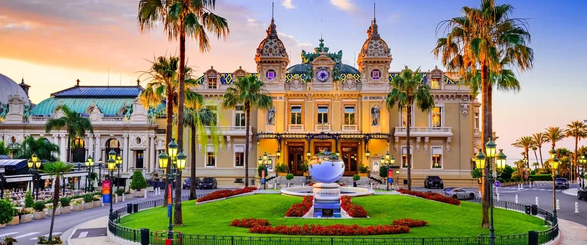 The historic Casino de Monte-Carlo building surrounded by palm trees, landscaped gardens, and a glowing sunset sky in Monaco.