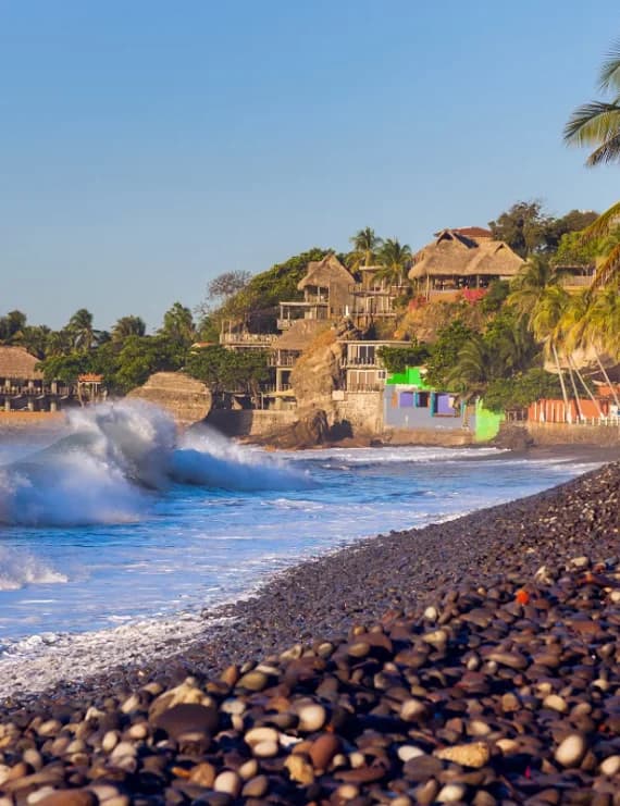 Powerful ocean waves crashing onto the black pebble shoreline with cliffside buildings at El Tunco beach.