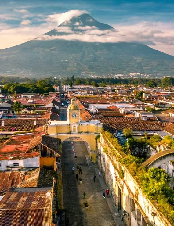 Aerial drone view of the iconic yellow Santa Catalina Arch, colonial rooftops, and Volcán de Agua in Antigua Guatemala.