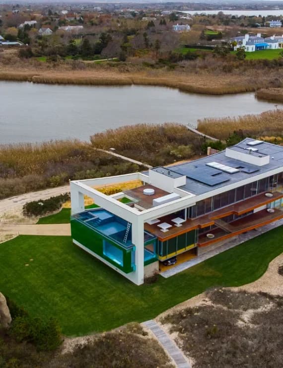 Aerial view of a modern glass luxury mansion with a rooftop pool and solar panels on a coastal bay in the Hamptons