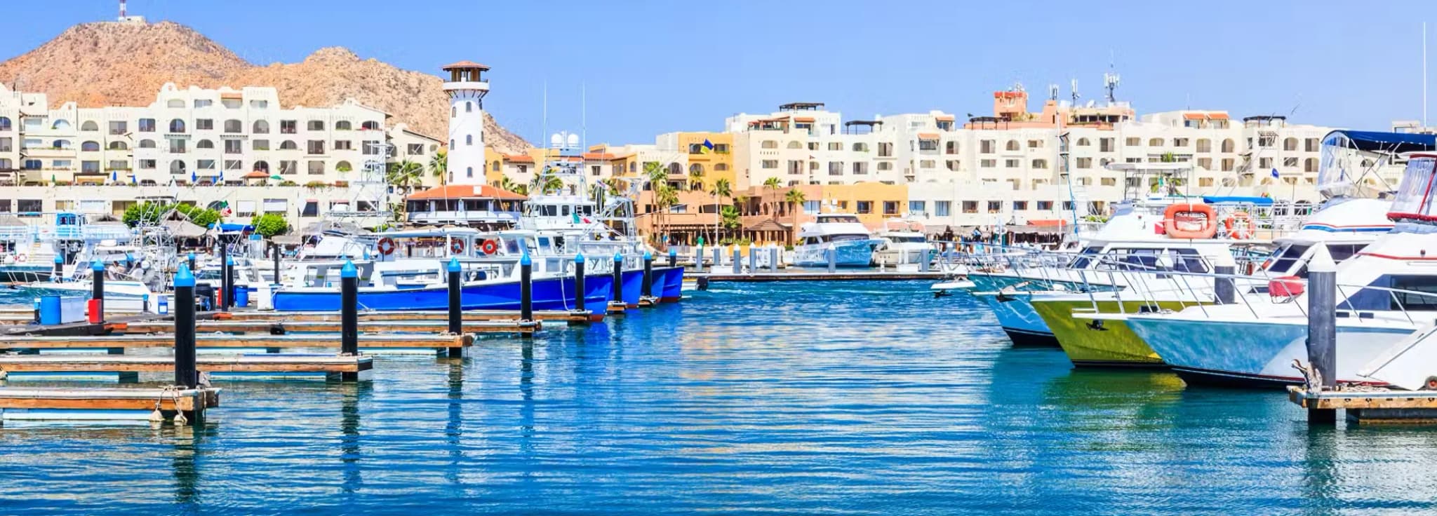 Marina Cabo San Lucas featuring luxury yachts, fishing boats, and the iconic lighthouse under a clear blue sky.