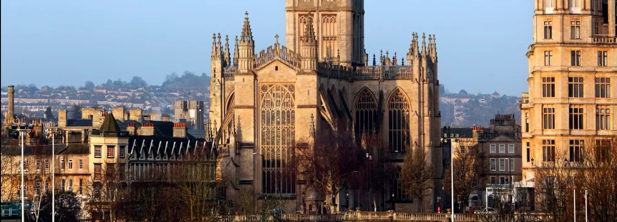 Exterior view of Bath Abbey showing its historic stone tower, stained glass windows, and the Union Jack flag flying under a clear sky in Somerset, England.