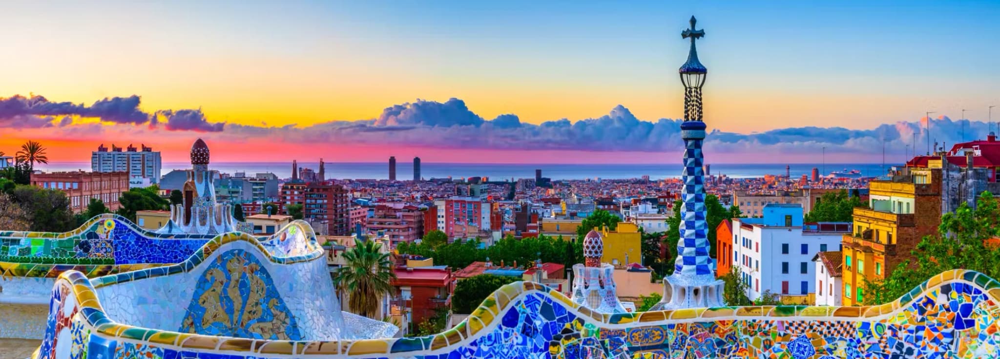 Colorful mosaic serpentine bench at Park Güell with a panoramic sunset view over Barcelona city skyline and the Mediterranean Sea in Spain.