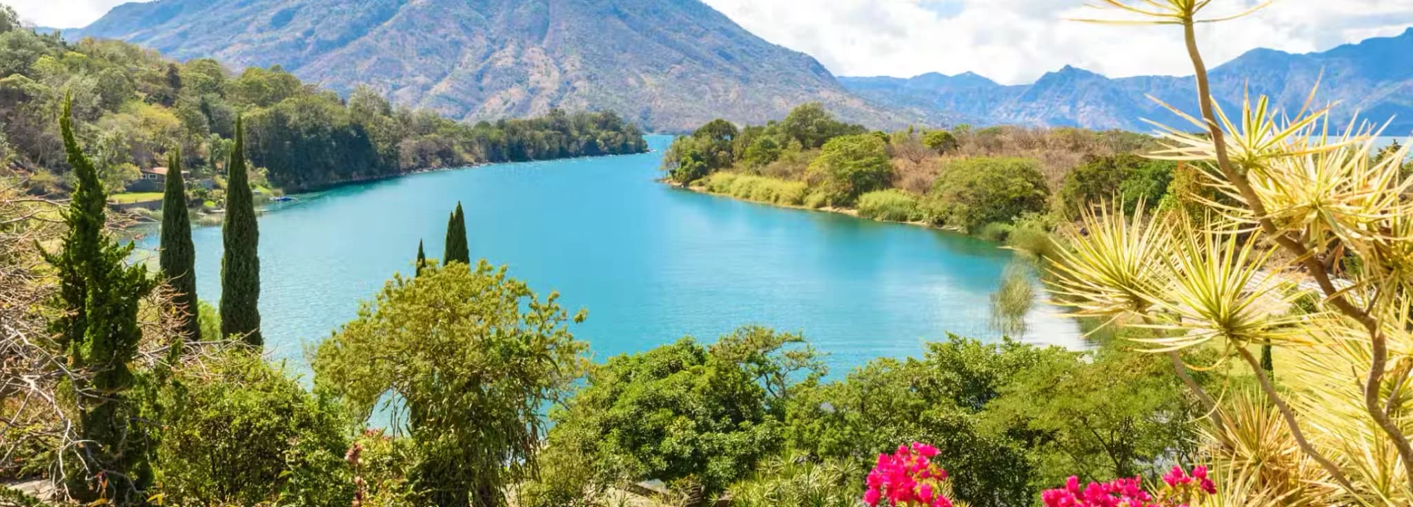Vibrant blue water of Lake Atitlán surrounded by lush green highlands, colorful flowers, and a majestic volcano in Guatemala.