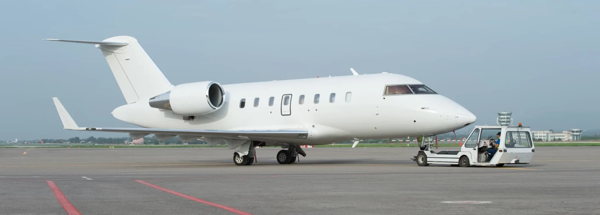 A white private jet aircraft being repositioned on the airfield tarmac by a small tow tractor.