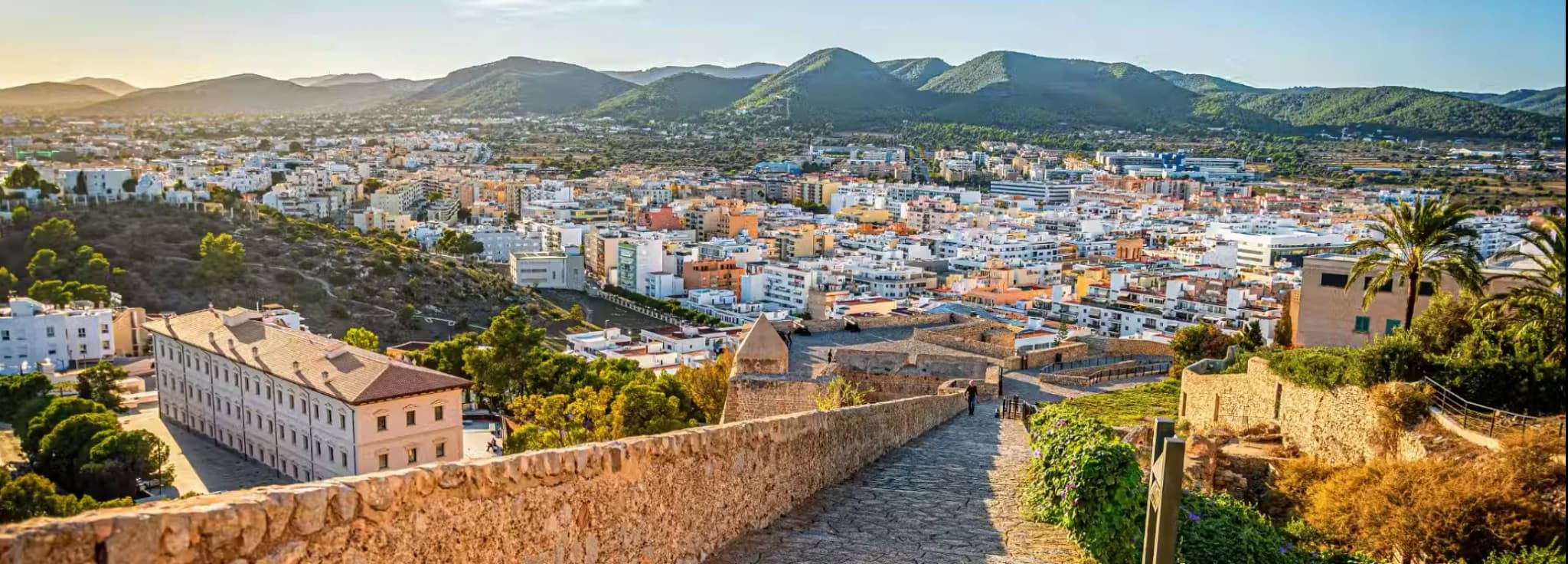 Historic stone pathway and ramparts of the Dalt Vila fortress overlooking the white buildings and hills of Ibiza Town, Spain.