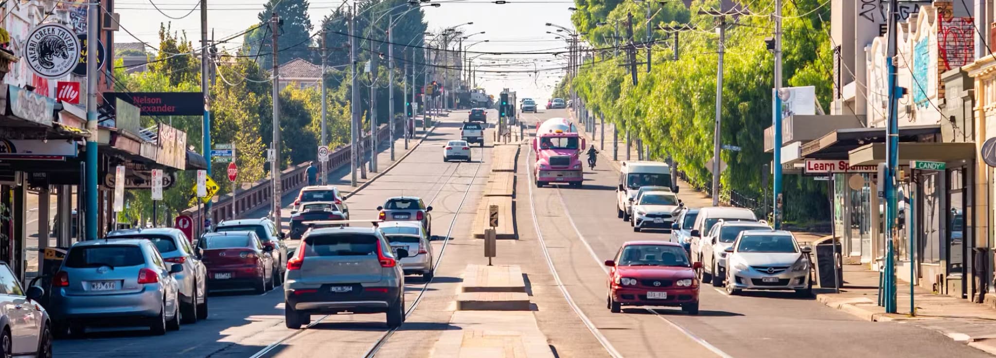 Street-level view of a bustling urban street with cars, trams, and storefronts under bright daylight.