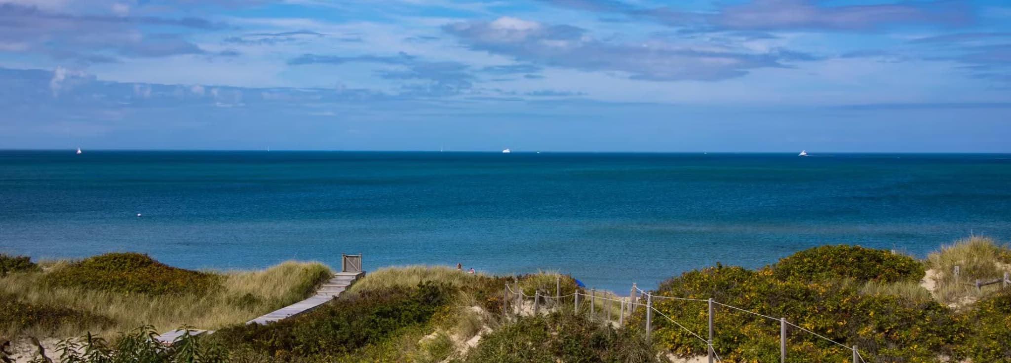 Wooden boardwalk walkway passing through grassy sand dunes leading to the blue ocean in Nantucket Massachusetts
