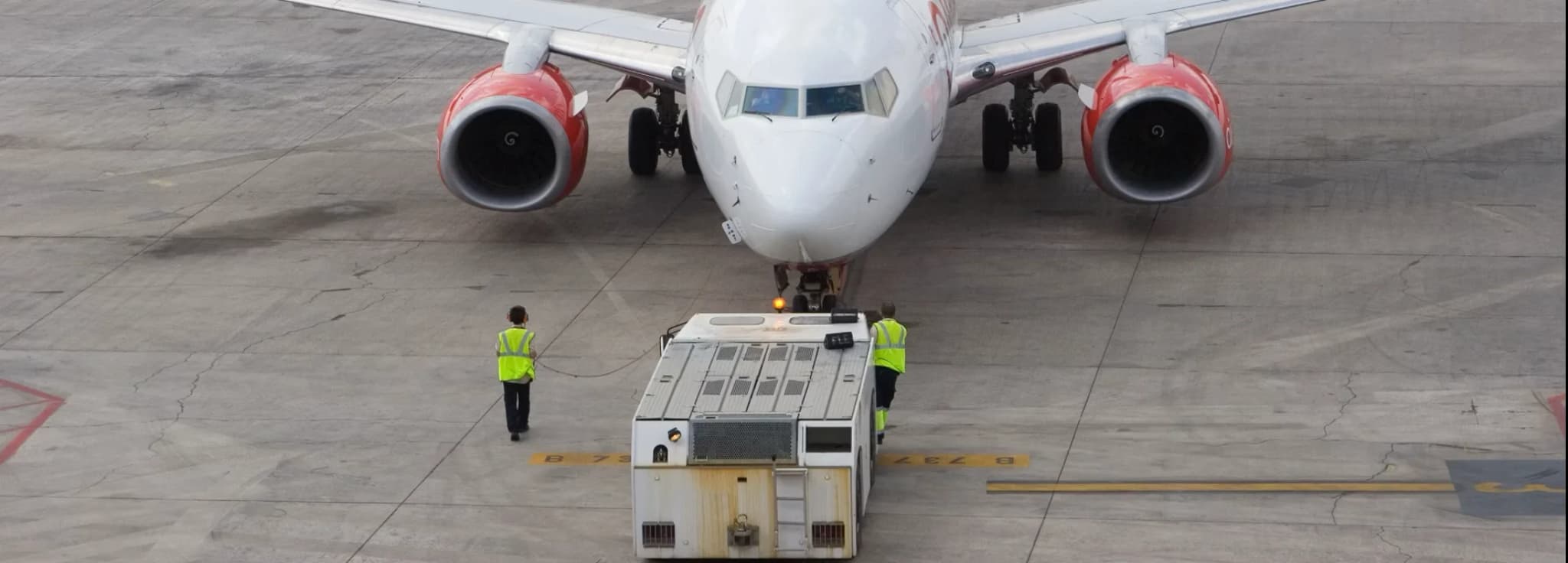 A commercial airliner parked on the concrete airport apron with a pushback tractor and ground crew in high-visibility vests positioned in front